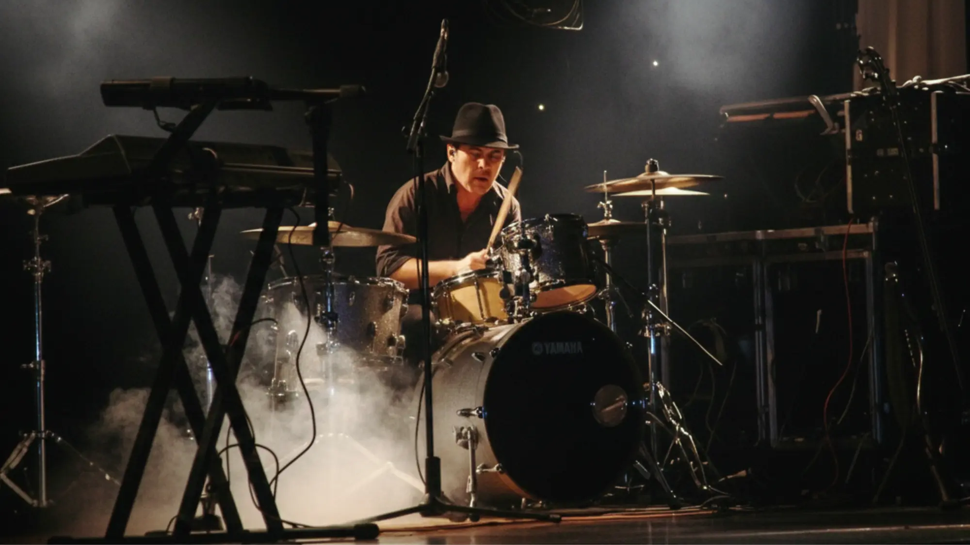 Simon playing a Yamaha kit on a dark stage in a trilby, lit through smoke, with keyboards and flight cases either side of him.