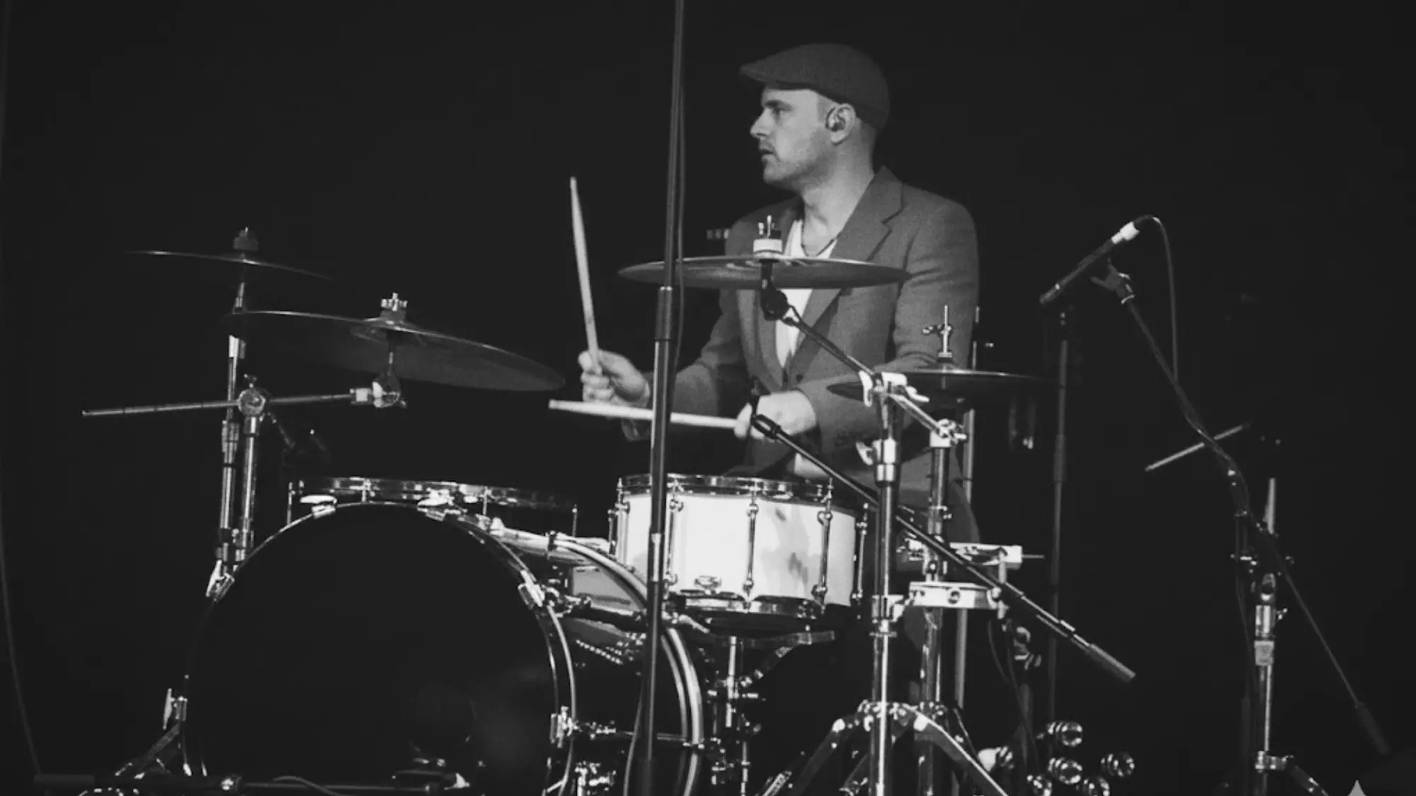 A black and white photograph of Simon mid-fill behind a drum kit, in a flat cap and a suit jacket, against a black stage.